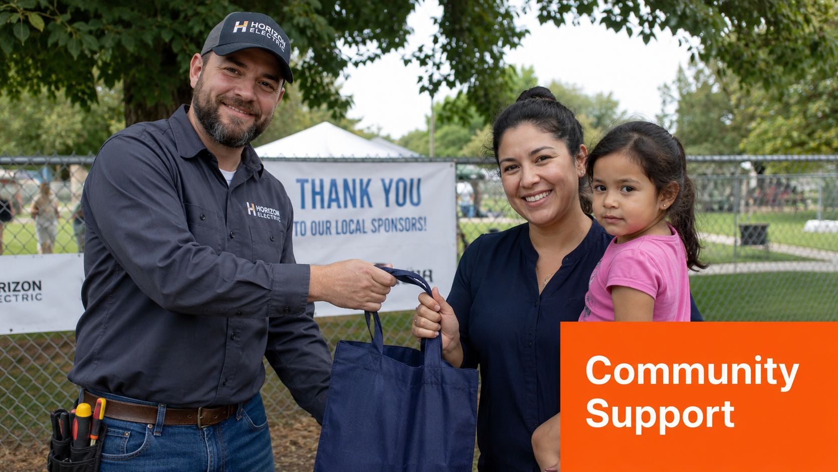 A smiling electrician in branded workwear hands a promotional bag to a woman holding her young daughter.